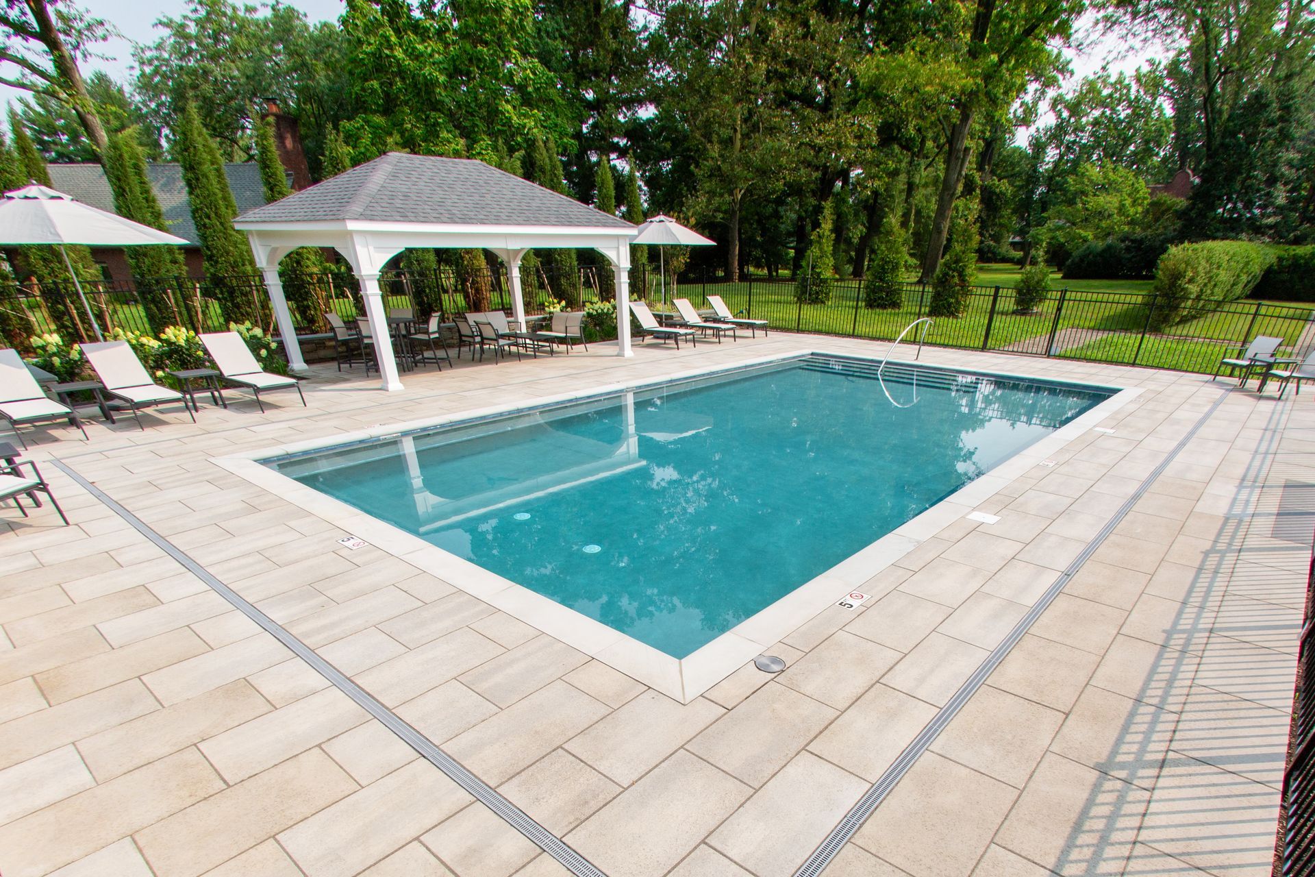Rectangular pool with beige patio, gazebo, lounge chairs, and trees in a backyard setting.