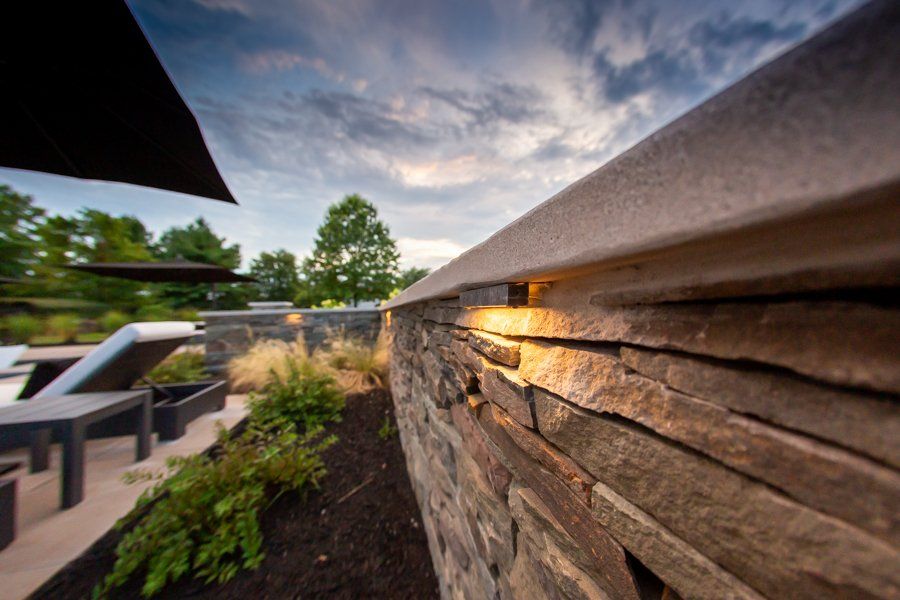 Stone wall with integrated lighting, overlooking a patio with lounge chairs, under a cloudy sky.