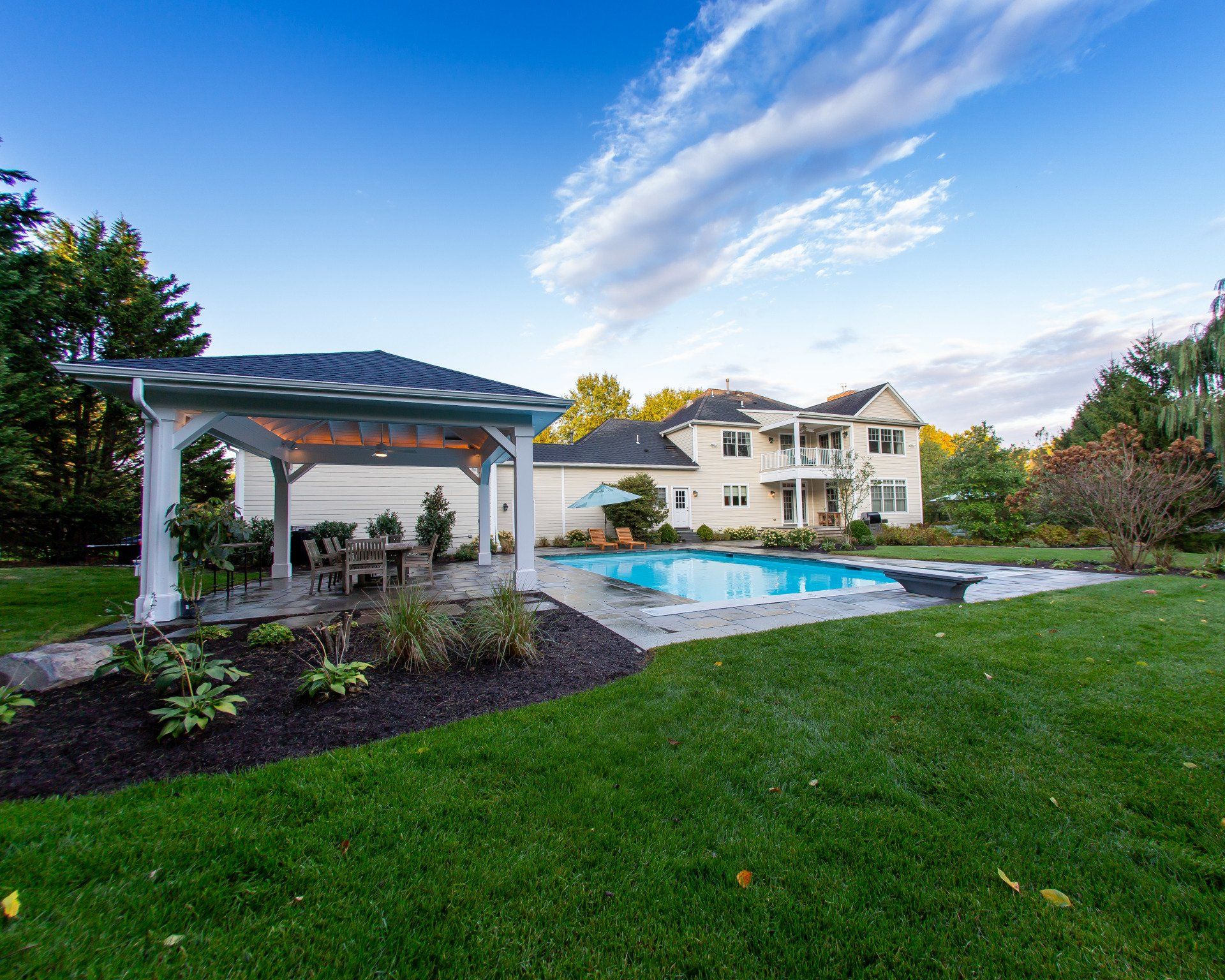 Backyard with pool, pergola, and large two-story house. Green lawn, blue sky with clouds.
