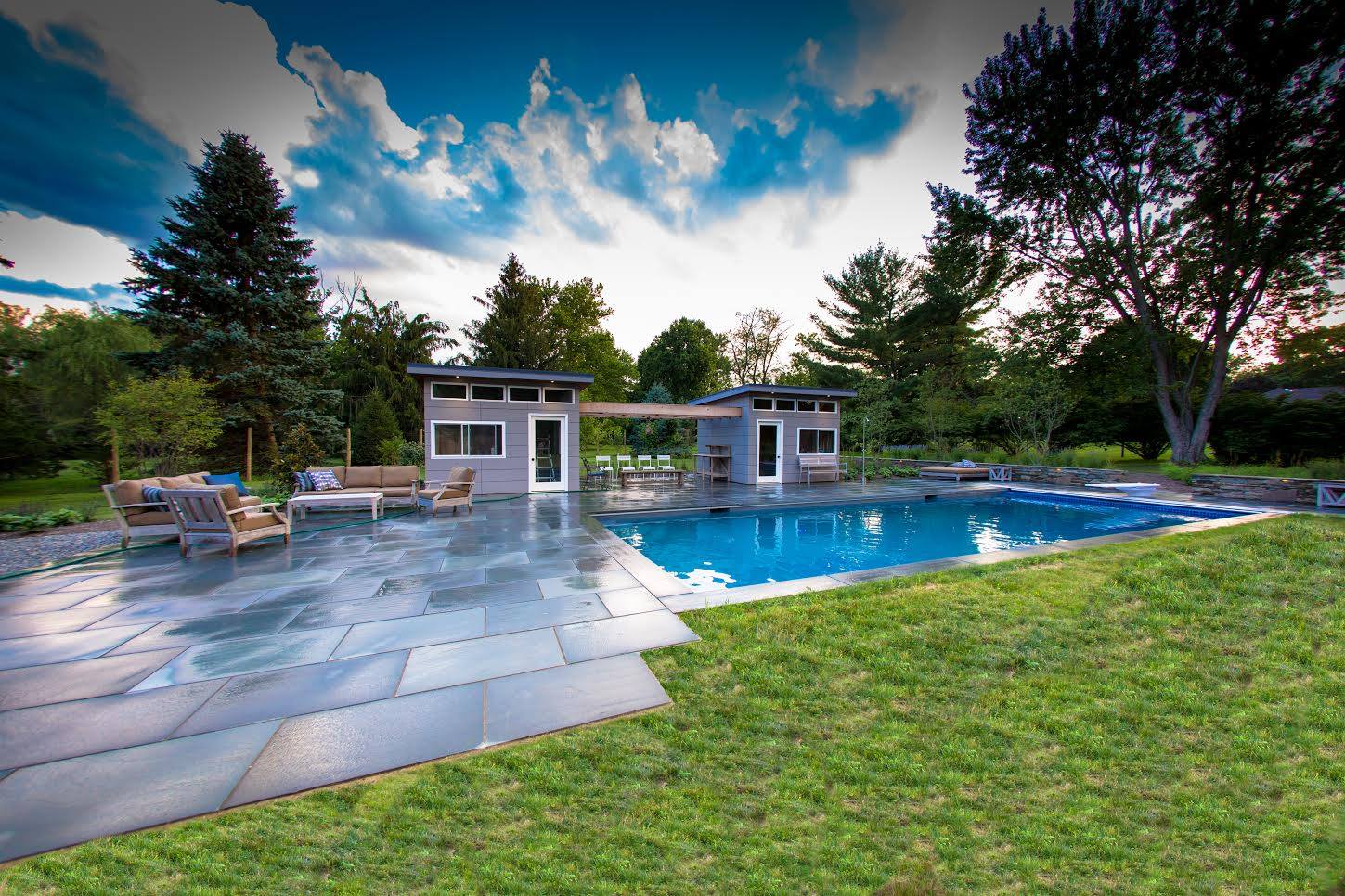 Poolside scene with pool, patios, small buildings, and lawn. Blue sky with clouds.