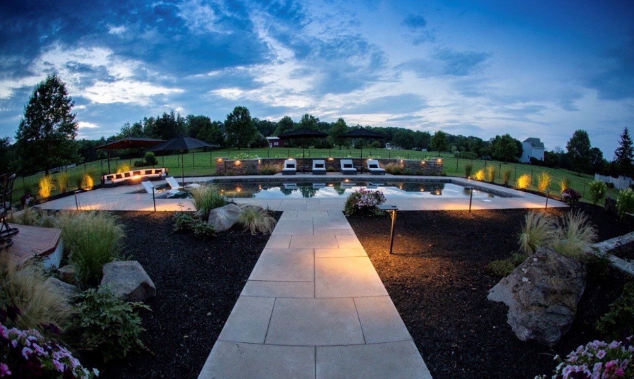 Night view of a pool and patio with landscape lighting and a cloudy sky.