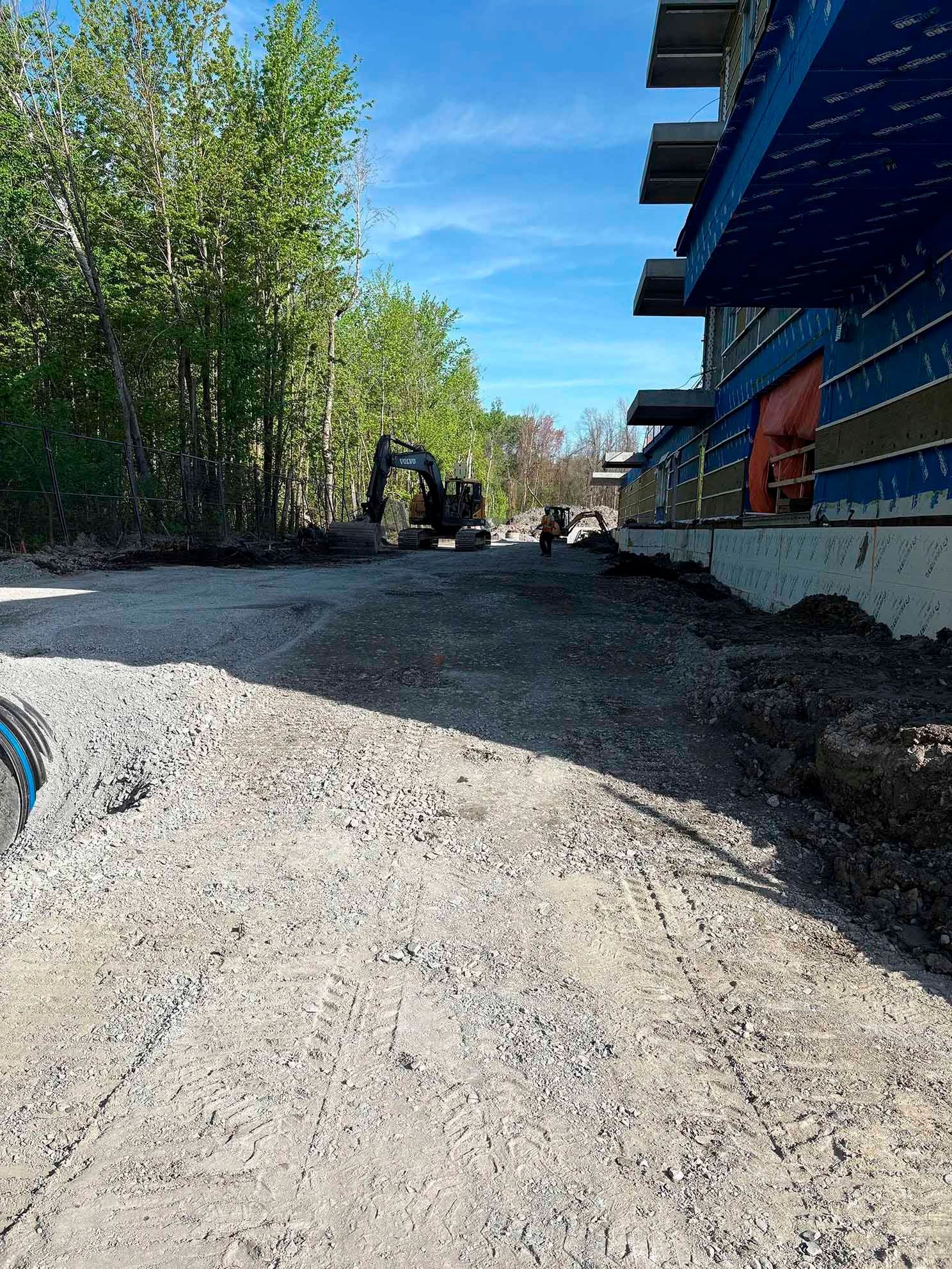 Zone de construction en gravier à côté du bâtiment. Pelleteuse et arbres en arrière-plan sous un ciel bleu.