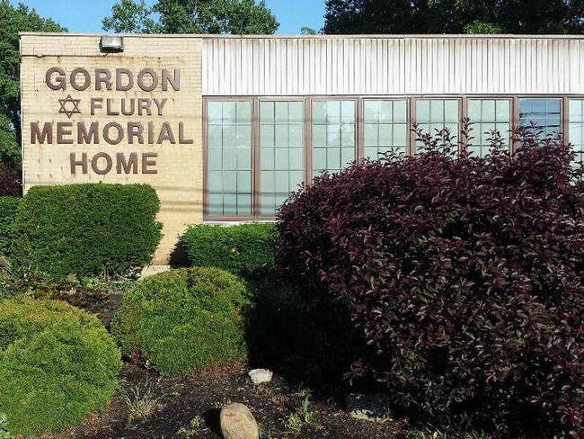 Gordon Flury Memorial Home sign on a white and beige building, with green and burgundy bushes in front.