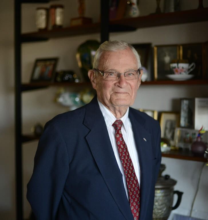Elderly man in a blue suit and red tie stands in front of a shelf with framed photos and objects.