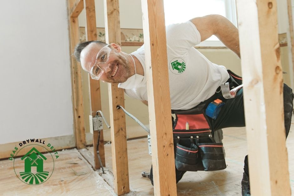 A drywall repair company worker in a white shirt is kneeling behind wooden wall studs, working on a residential construction project. The contractor performs renovation work near Marmont St, and Quadling Ave, Coquitlam, BC.