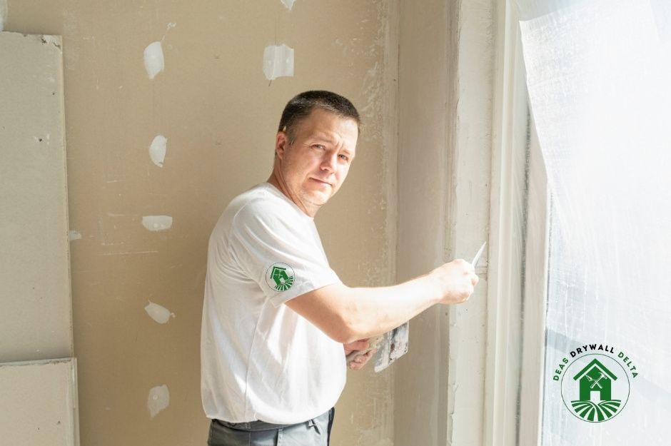 A drywall repair company professional in a white shirt smiles while applying joint compound to drywall using a putty knife. He is working in a residential construction site with visible drywall patches, providing quality finishing services near Edinburgh St, and 22nd St, New Westminster, BC.