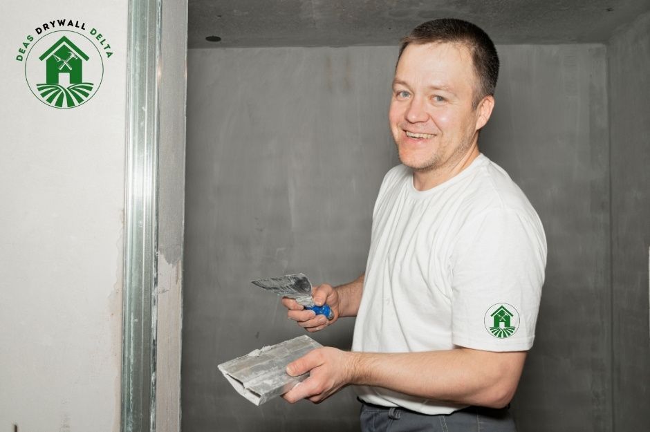 A drywall repair company professional in a white shirt smiling while holding a putty knife, standing next to freshly applied drywall compound on a wall in a residential construction site near 193 St, and 118b Ave, Pitt Meadows, BC.