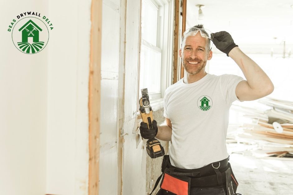 A drywall repair company worker, a smiling man in a white shirt and a tool belt, holds a power drill while standing in a commercial construction site with exposed wooden framing and drywall sheets visible in the background near Cornwall Dr, and No 1 Rd, Richmond, BC.