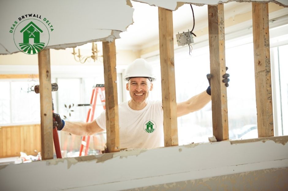 A drywall repair company worker in a white hard hat and tank top stands with arms raised in an unfinished interior space with exposed wooden wall studs and bright lighting, working on a commercial drywall installation near Carnarvon St and W 55th Ave, South Vancouver, BC.