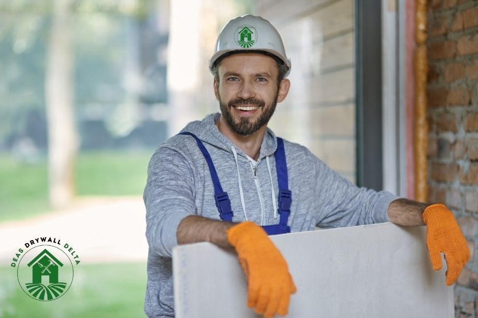A drywall repair company worker, a smiling bearded man wearing a white hard hat and blue overalls, leans against white drywall with his arms resting on the surface while wearing orange work gloves in a residential construction setting near 70a Ave, and 201b St, Willoughby, BC.