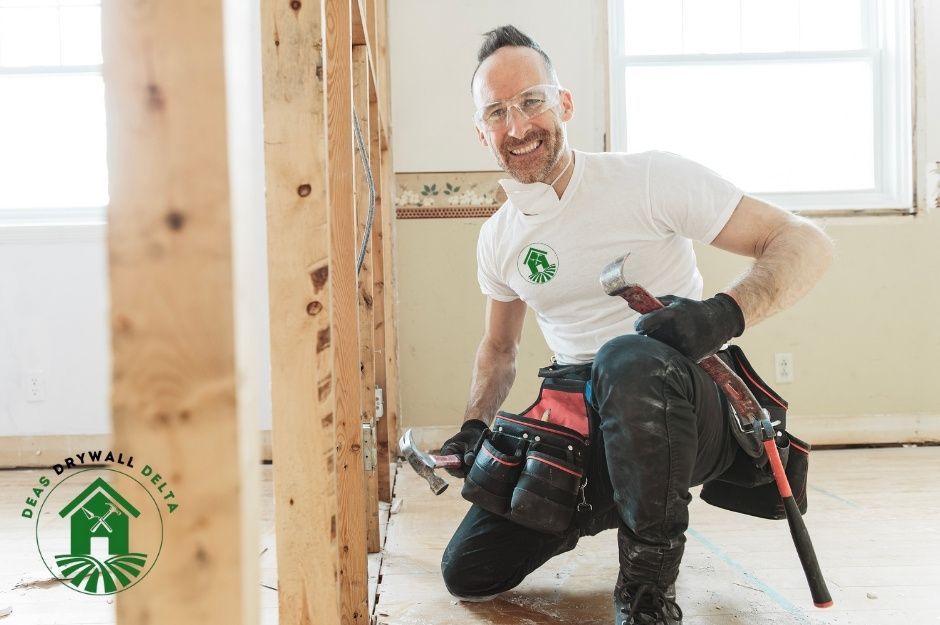A drywall repair contractor kneels on a commercial construction site floor, smiling while wearing work clothes and a tool belt. He holds a hammer in his hand, with wooden framing visible behind him in the unfinished room near 2nd St, and 2nd Ave, Port Moody, BC.