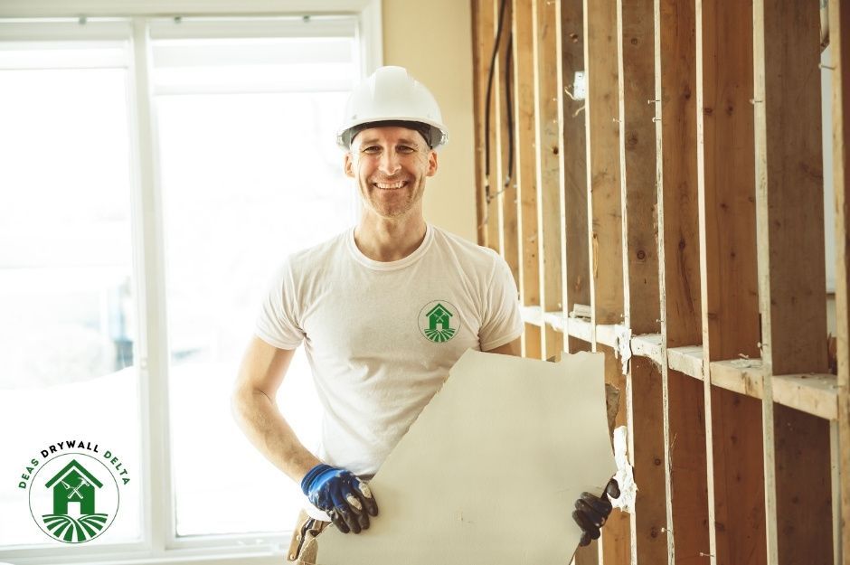 A drywall repair contractor is wearing a white hard hat and t-shirt, smiling while holding drywall sheets in a residential construction site with exposed wooden wall framing and natural lighting from windows in the background near Blackwood St, and Prospect Ave, White Rock, BC.