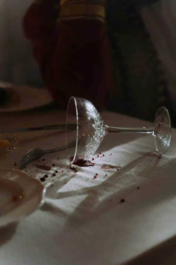 A crystal wine glass lies on its side on a white tablecloth, surrounded by red wine stains and splatters.