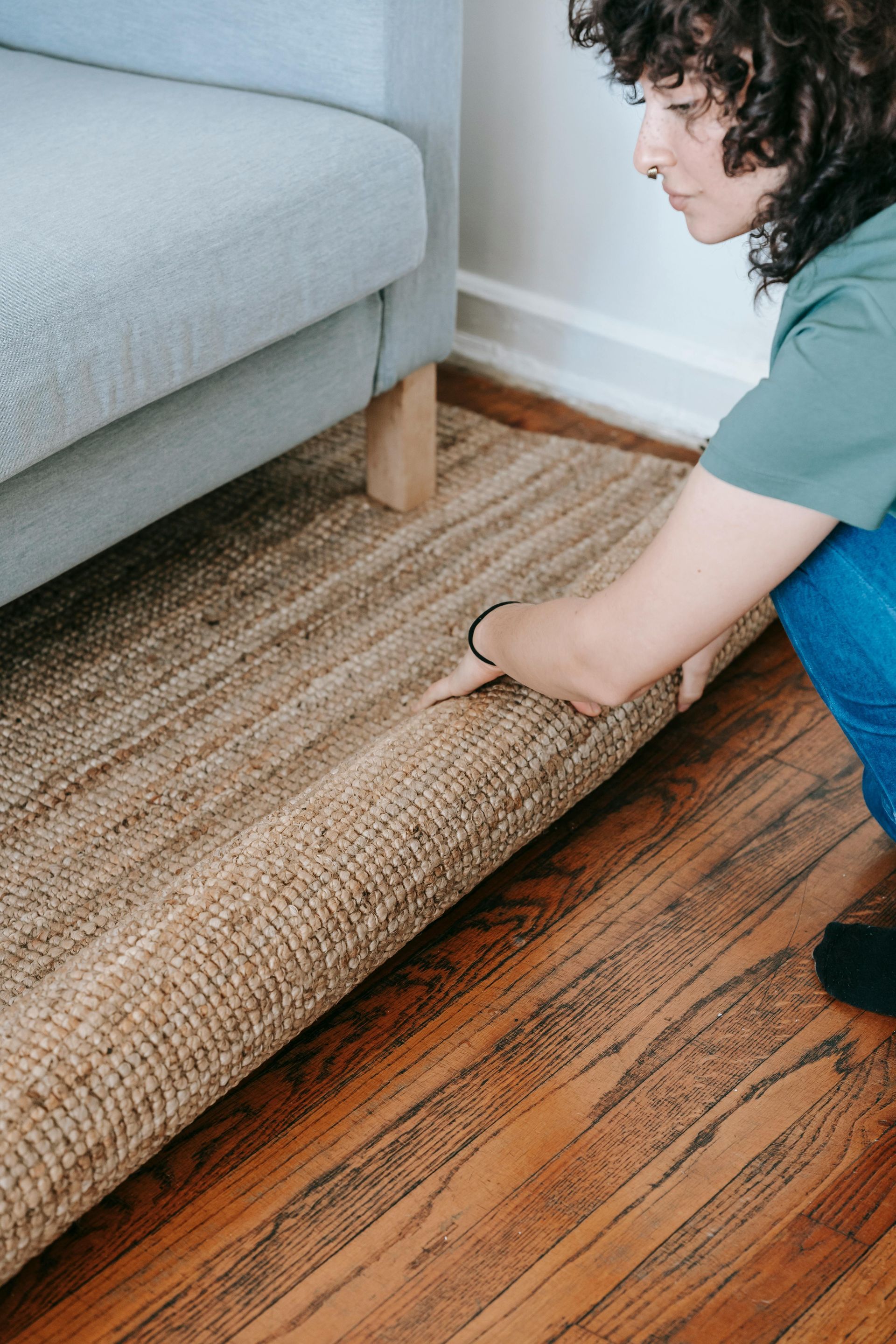 A man unrolls a patterned rug on a wooden floor, while a woman in casual clothes works on a laptop on a bed. 