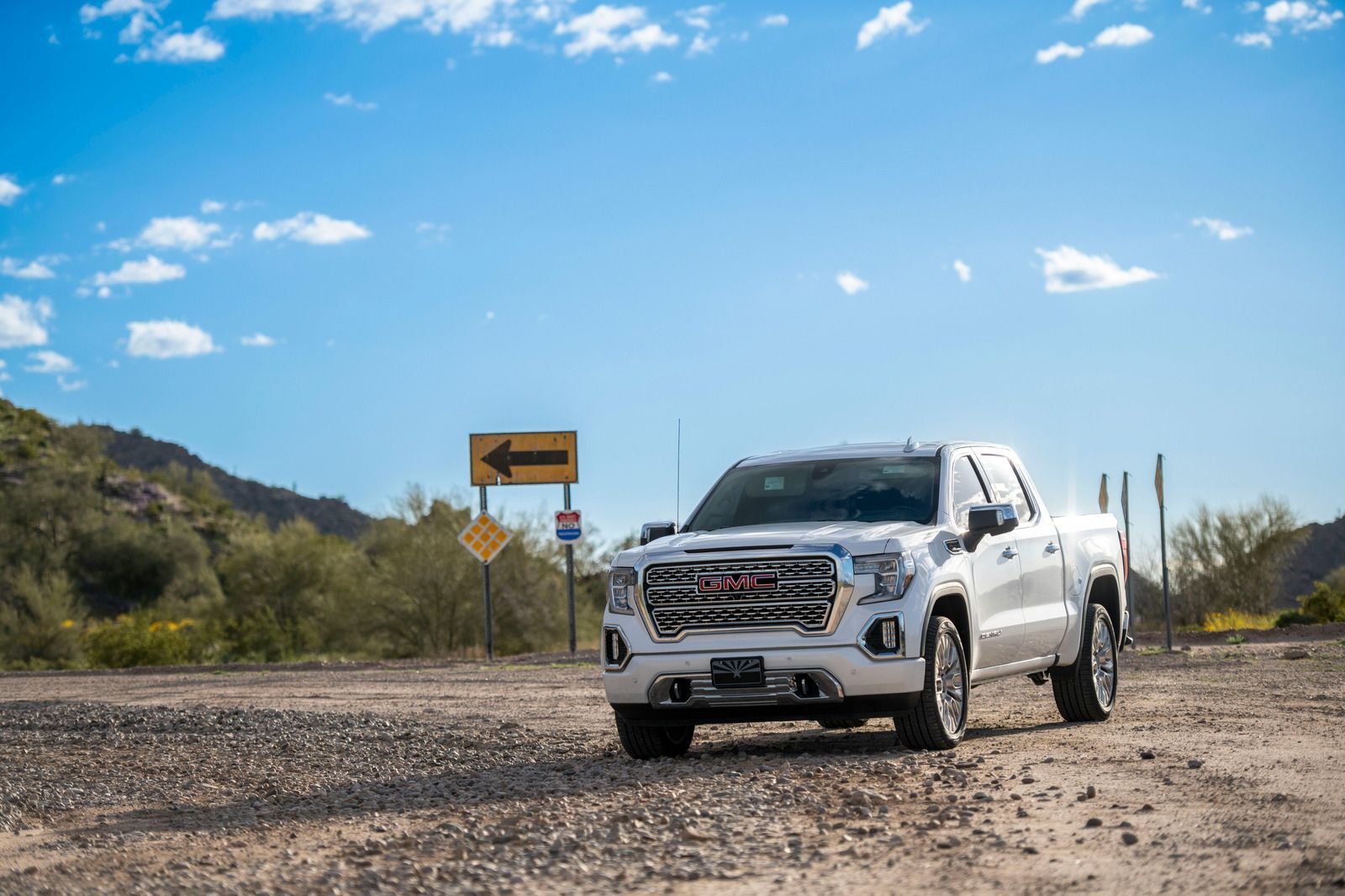 White GMC truck on a dirt road, with mountain backdrop and a blue sky.