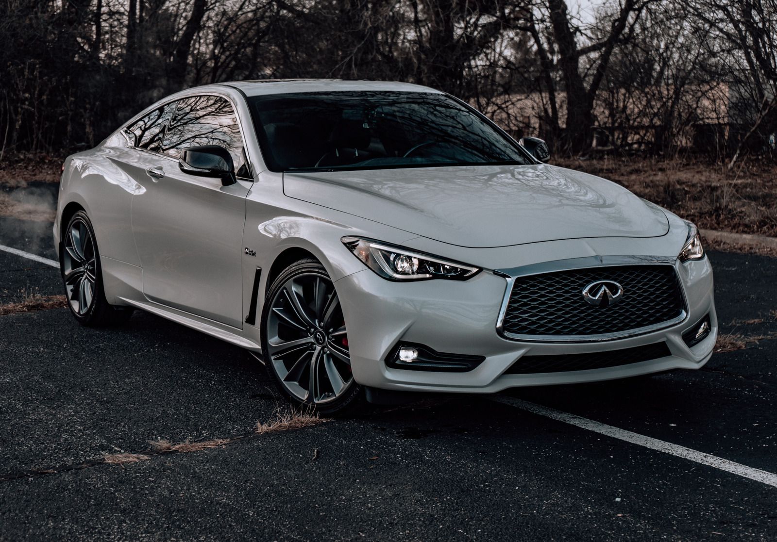 White Infiniti Q60 coupe on a dark road; dark wheels, sporty design, trees in the background.