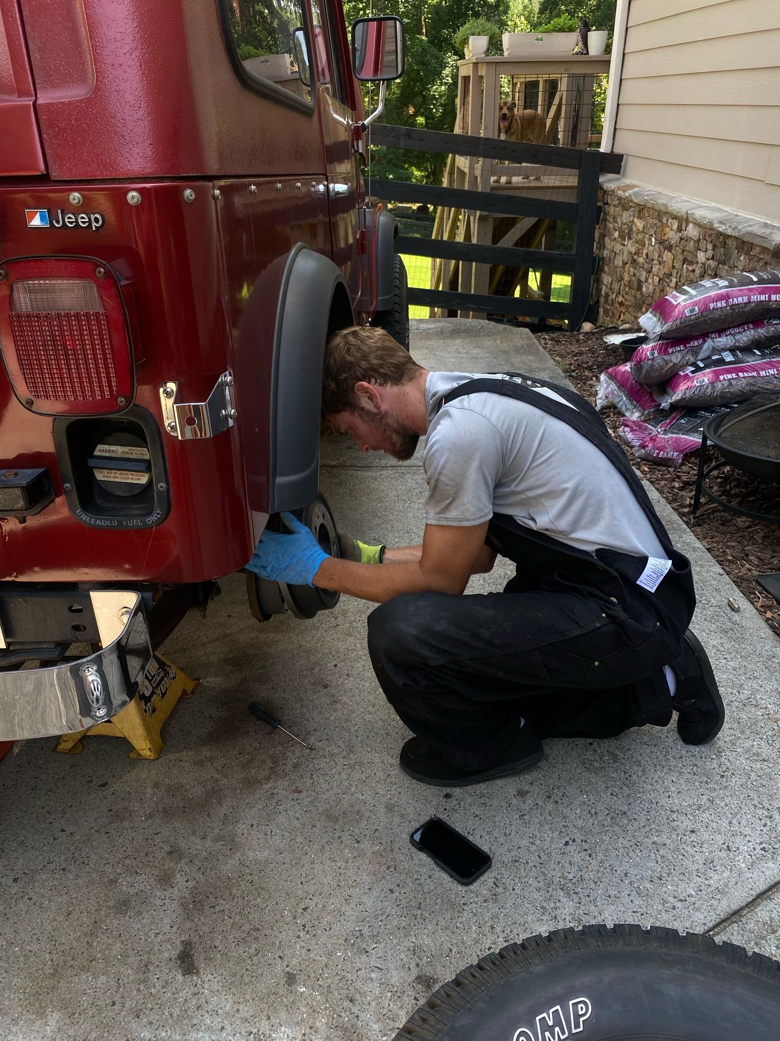 Man in overalls working on a red Jeep's rear tire. He's wearing gloves, squatting in a driveway.