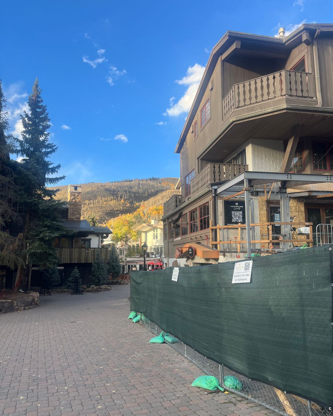 Building under construction, green fence, brown and beige exterior, street, mountain background, daytime.