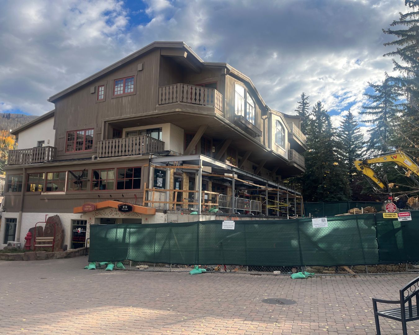 Building under construction with green fencing and a yellow excavator, brown exterior.