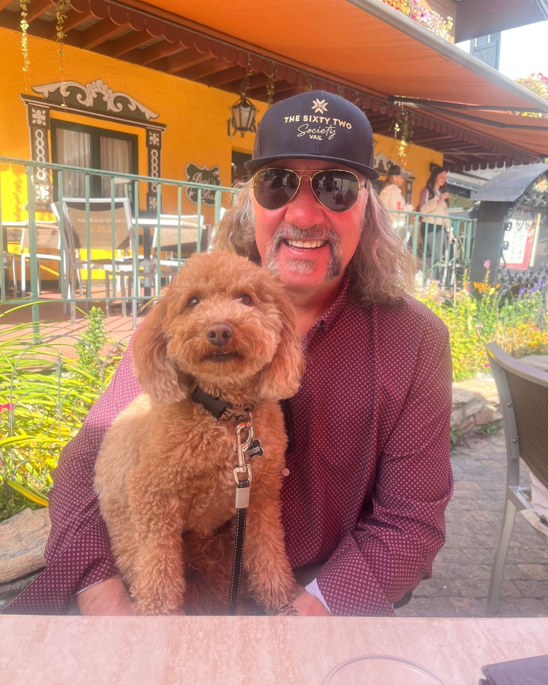Man with sunglasses and a dog in his lap, sitting outside.