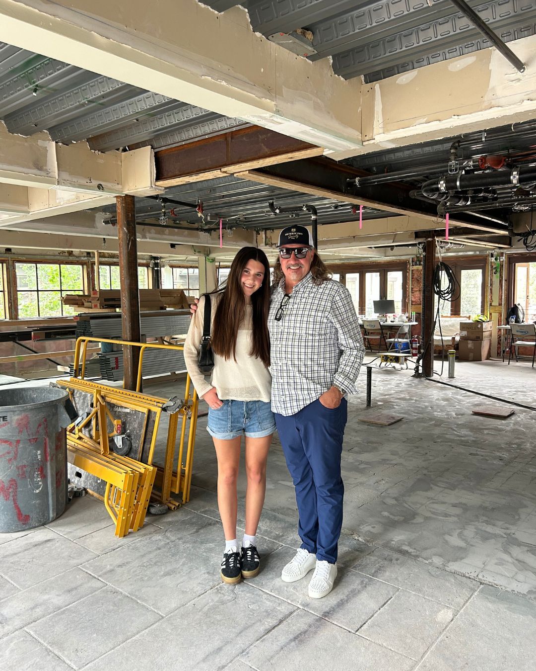 Two people pose in a construction site. Girl with long hair, boy in cap. Interior with exposed ceiling and debris.