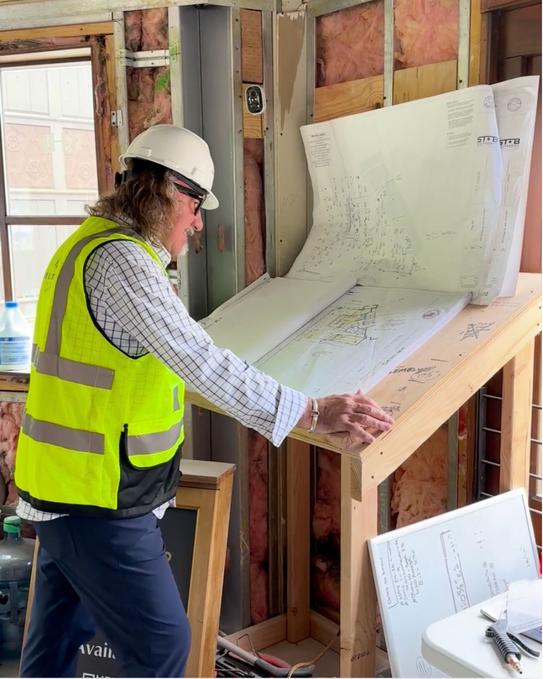 Person in hardhat and vest examines blueprints on a wooden drafting table at a construction site.