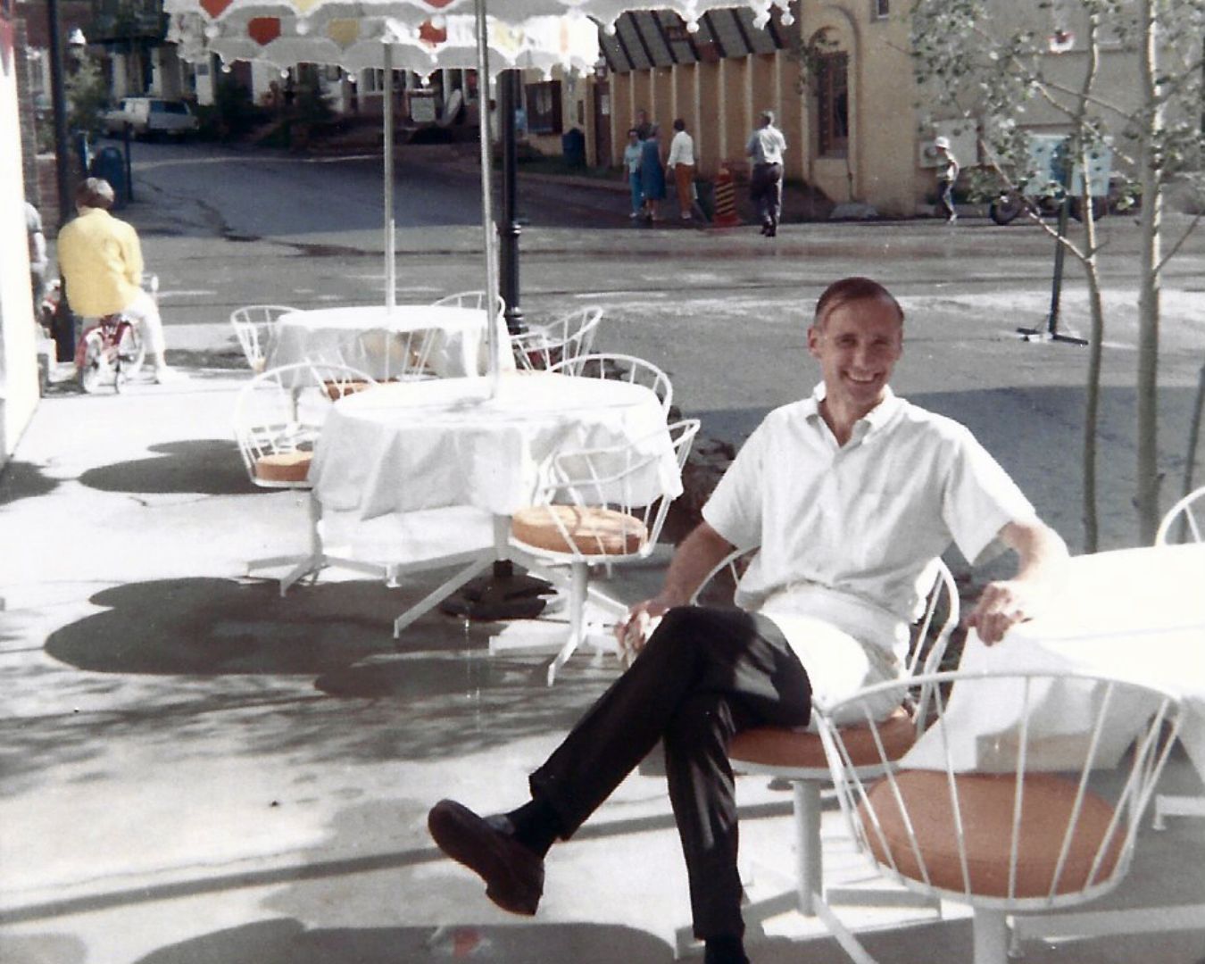 Man in white shirt and dark pants seated at outdoor table, smiling.