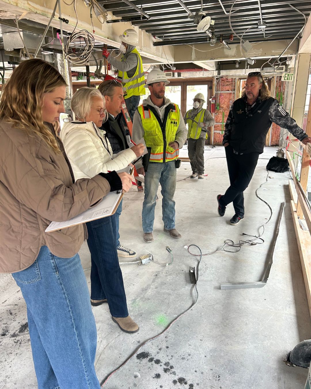 People in construction site discussing; workers in background; interior with exposed ceiling, wires, and building materials.