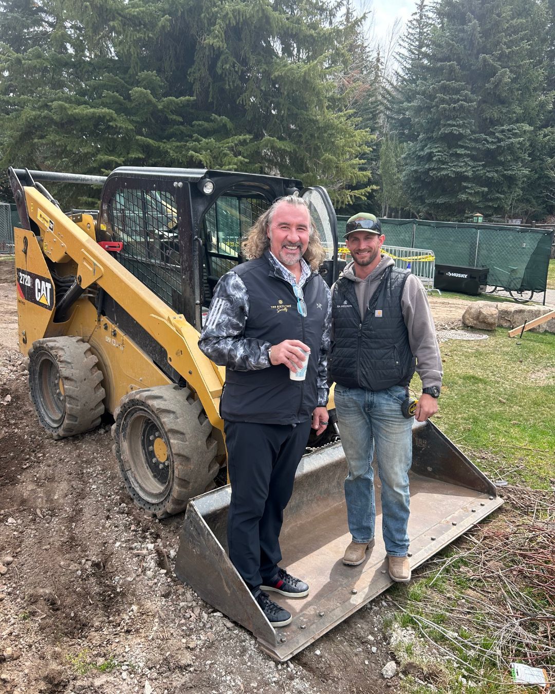 Two men beside a yellow CAT skid steer loader. One drinks coffee, the other smiles. Outdoor setting.