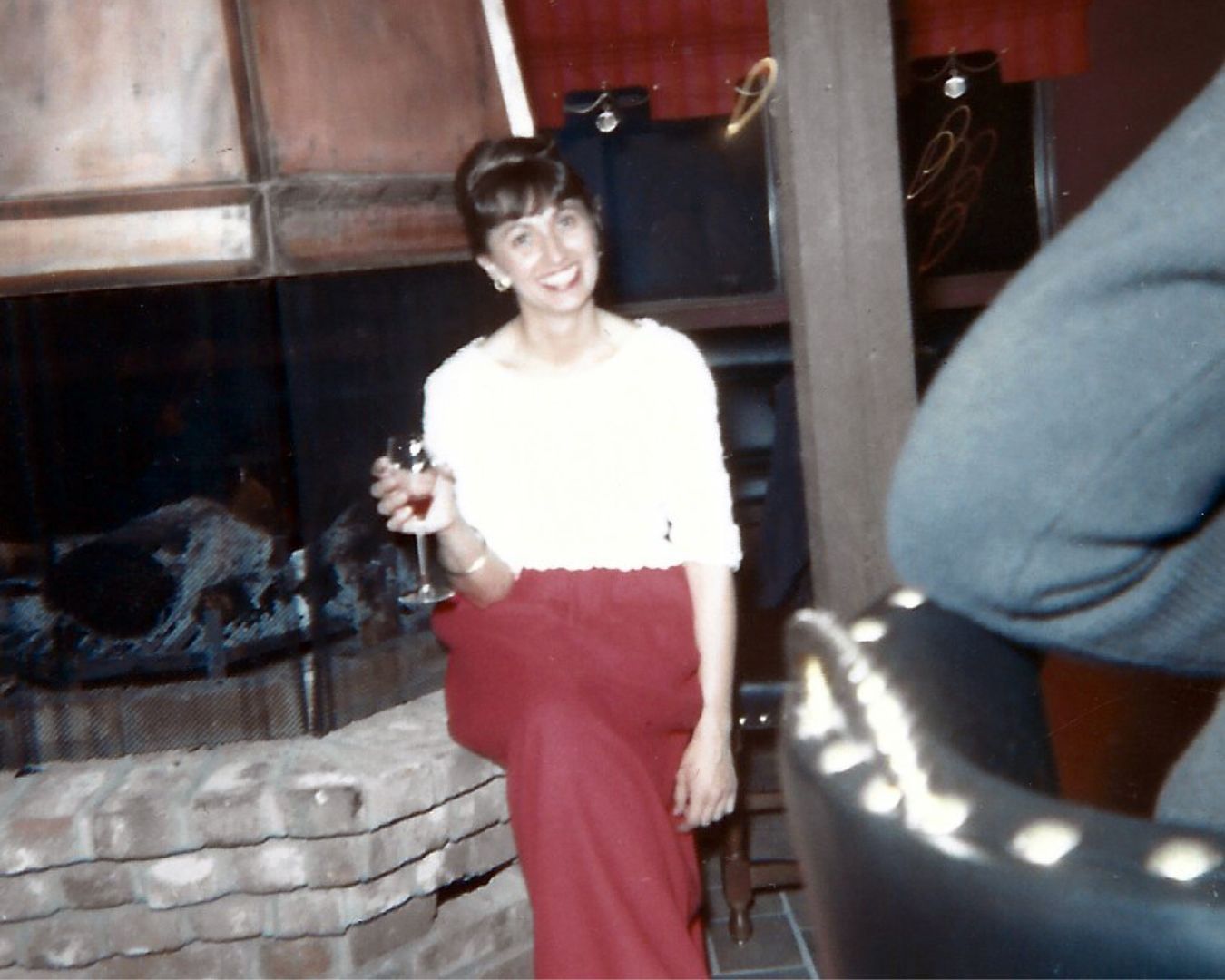 Woman in red pants and white top smiling, holding wine glass, by a fireplace.