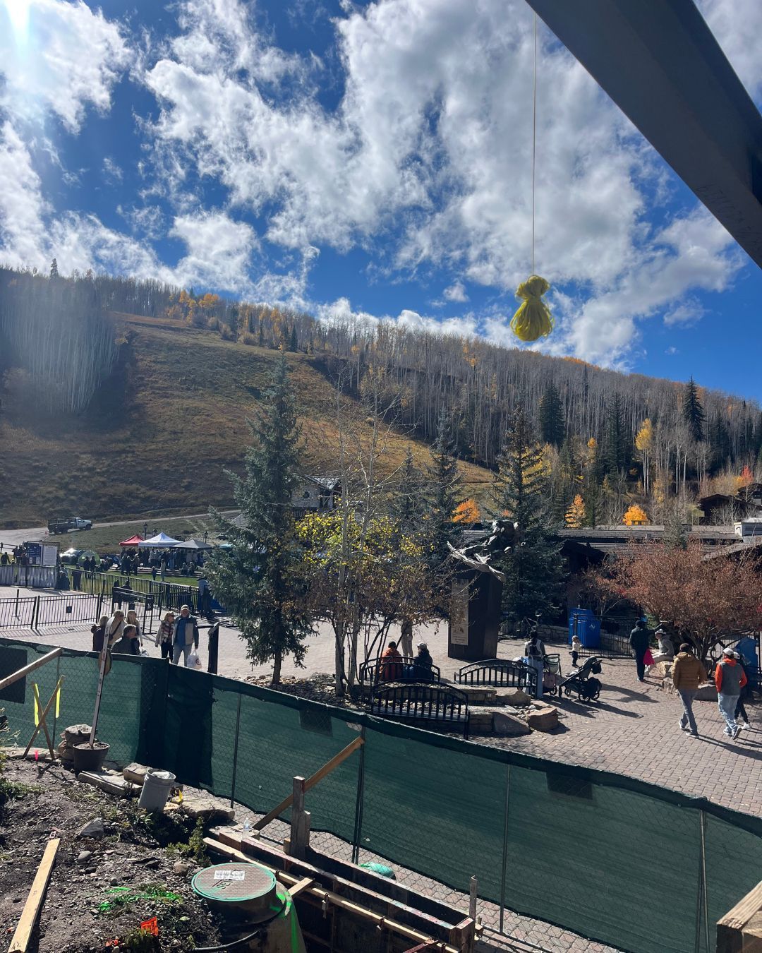 People gather outdoors near a ski slope on a sunny day; a yellow balloon floats above.