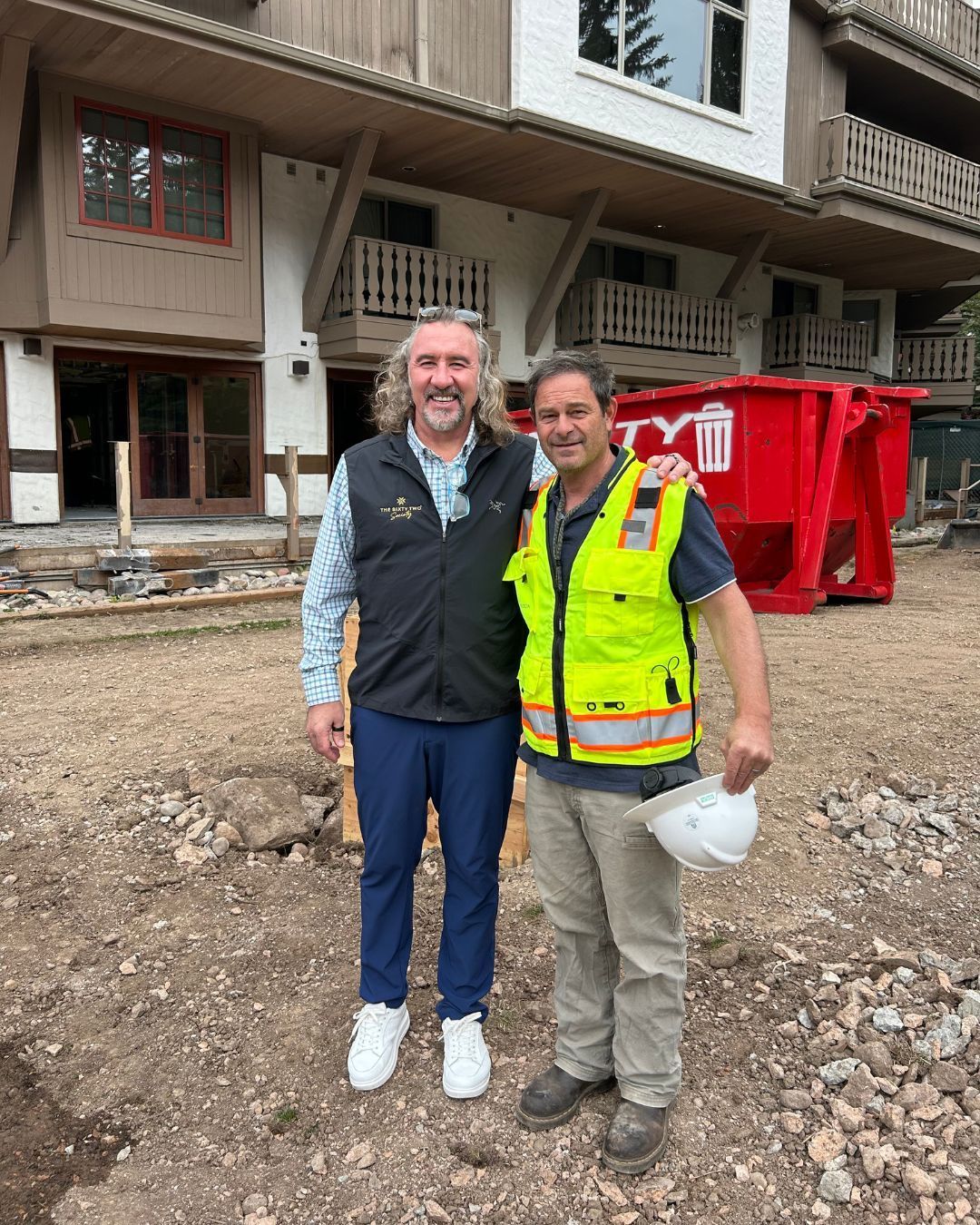 Two men posing outside a building under construction. One in a vest, one in a reflective vest.