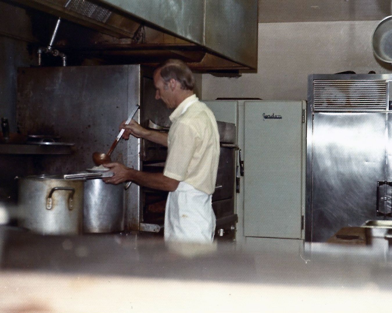 Man in a kitchen, wearing apron, ladling food from a large pot. Stainless steel appliances and a hood.