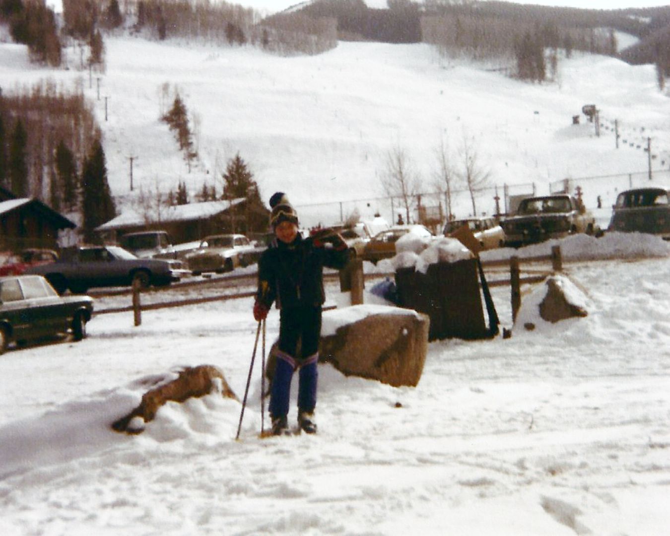 Person stands in snow with skis, lift and cars in the background.