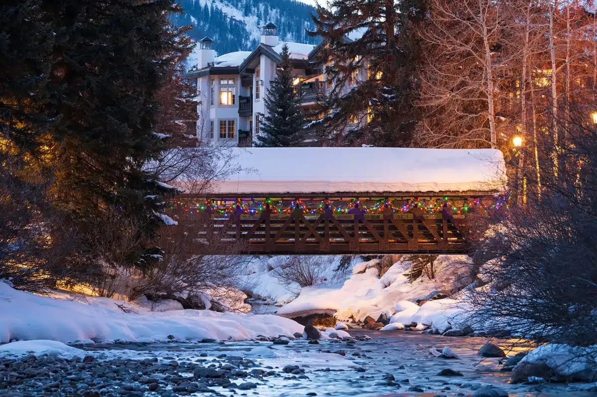 Snow-covered bridge with colorful lights over a flowing river. A building is visible in the background.