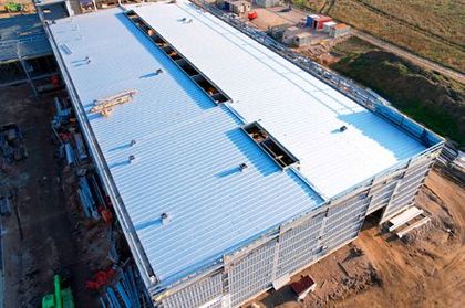 Aerial view of a large, unfinished industrial building with a gray metal roof under construction.