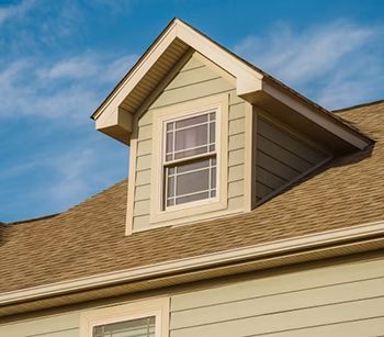 Dormer window on a house with light green siding, tan roof, and a clear blue sky.