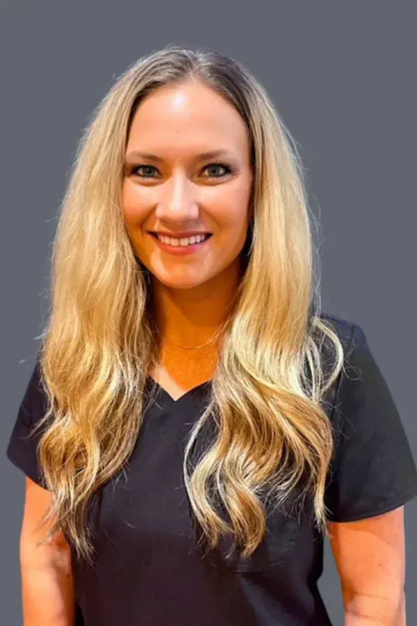 A woman with long blonde hair is wearing a black scrub top and smiling.