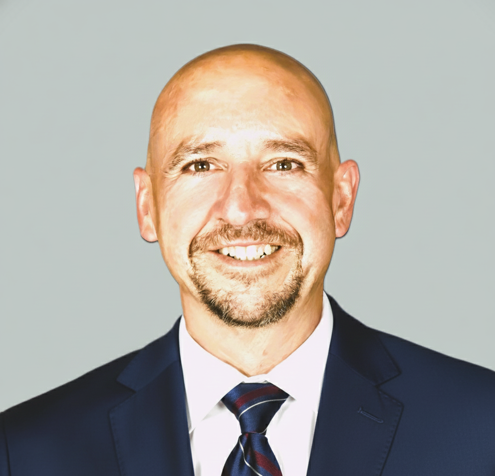 Man in a blue suit smiles, looking at the camera. He has a shaved head, goatee, and a patterned tie.