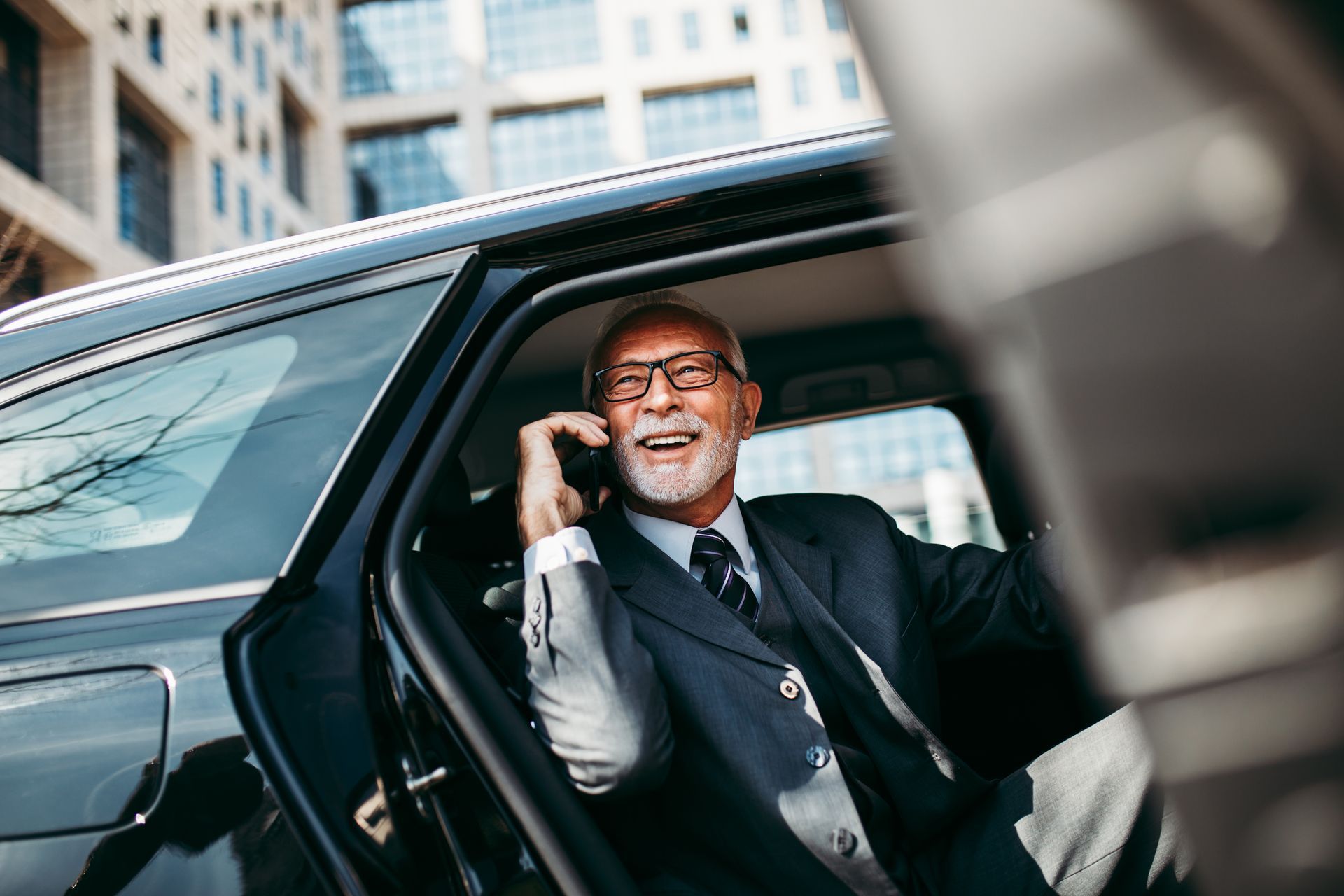 Man in a suit smiles while talking on phone in a car, door open, city background.