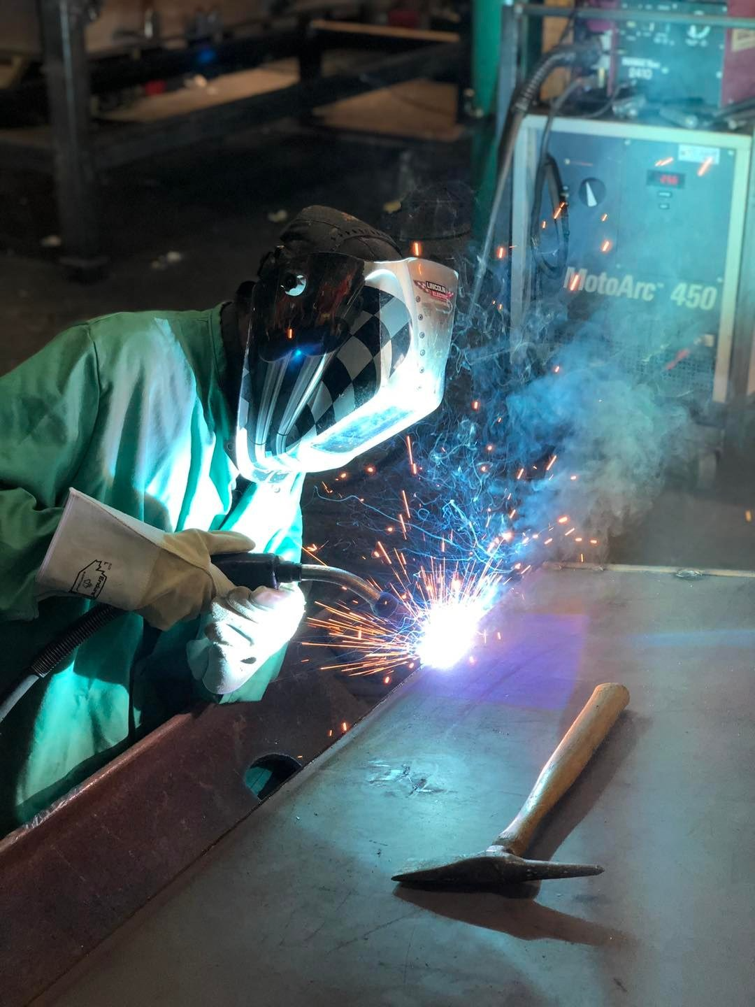 Un soudeur en chemise turquoise et visière de protection soude du métal ; des étincelles jaillissent dans l’atelier.