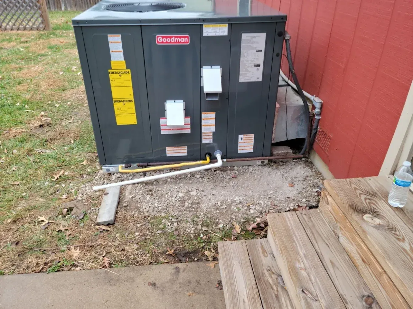 Outdoor HVAC unit beside a red wall, with yellow electrical conduit and wooden steps in the foreground