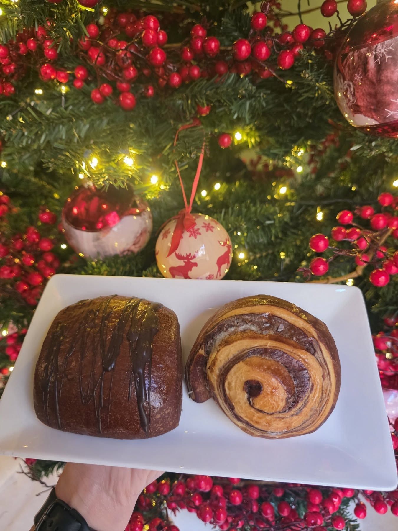 Due pasticcini su un piatto bianco, tenuti davanti a un albero di Natale decorato con luci e ornamenti rossi.