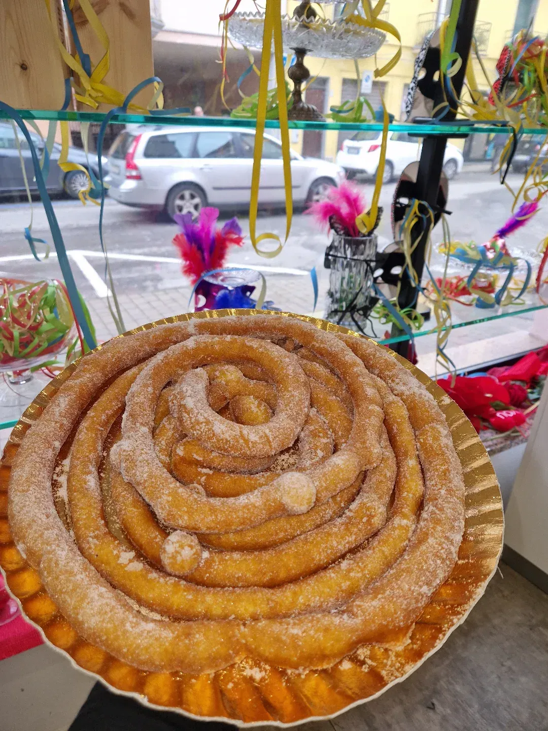 Grande pasta sfoglia fritta a forma di spirale, spolverata di zucchero, esposta su un piatto d'oro nella vetrina di un negozio.