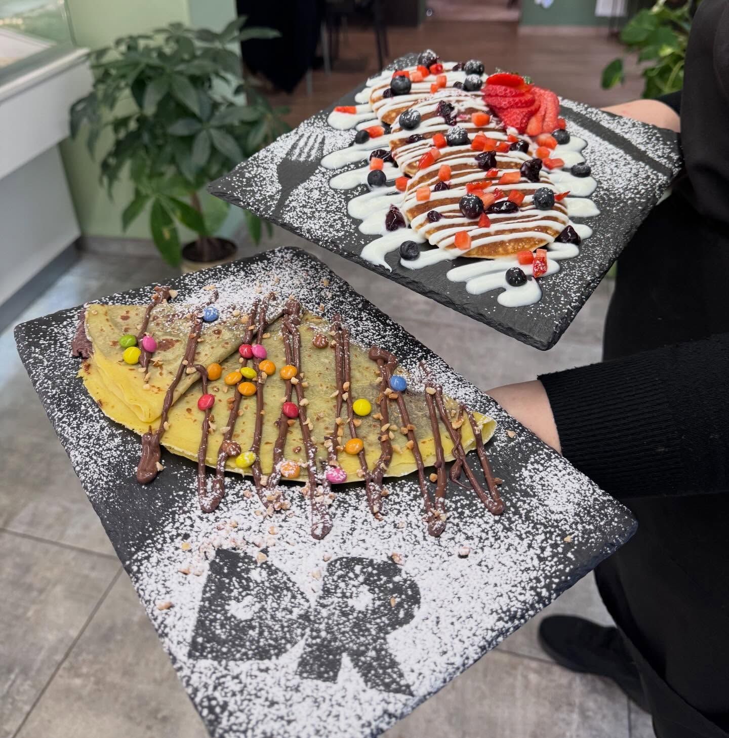 Crêpes guarnite con caramelle, frutti di bosco e cioccolato su vassoi di ardesia nera, tenuti da una persona.