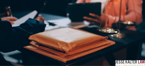 Close-up photograph of orange manila folders and white documents on a dark surface, with blurred figures in business attire