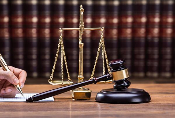 A gold scales of justice and dark wood-handled gavel rest on a wooden desk next to an inkwell and pen, with a blurred