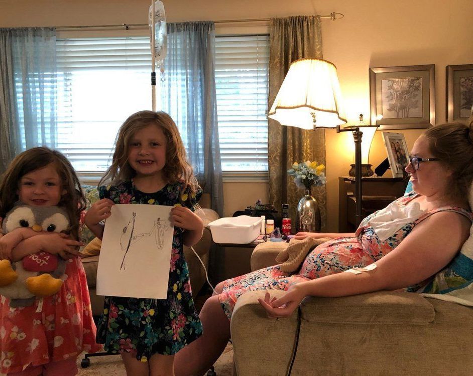 Two little girls are standing next to a pregnant woman in a living room