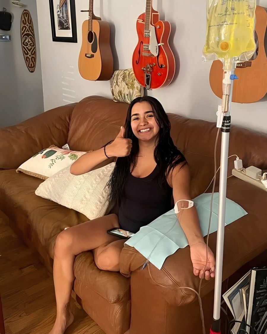 Smiling young woman gives a thumbs up while receiving an IV infusion at home, sitting on a leather couch with guitars on the wall.