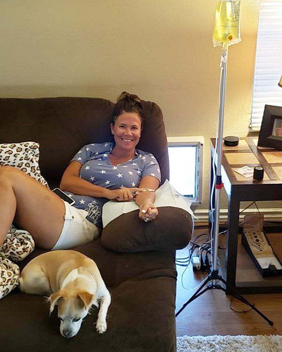 Smiling woman sitting comfortably on a couch with her small dog nearby, receiving an at-home IV therapy infusion.